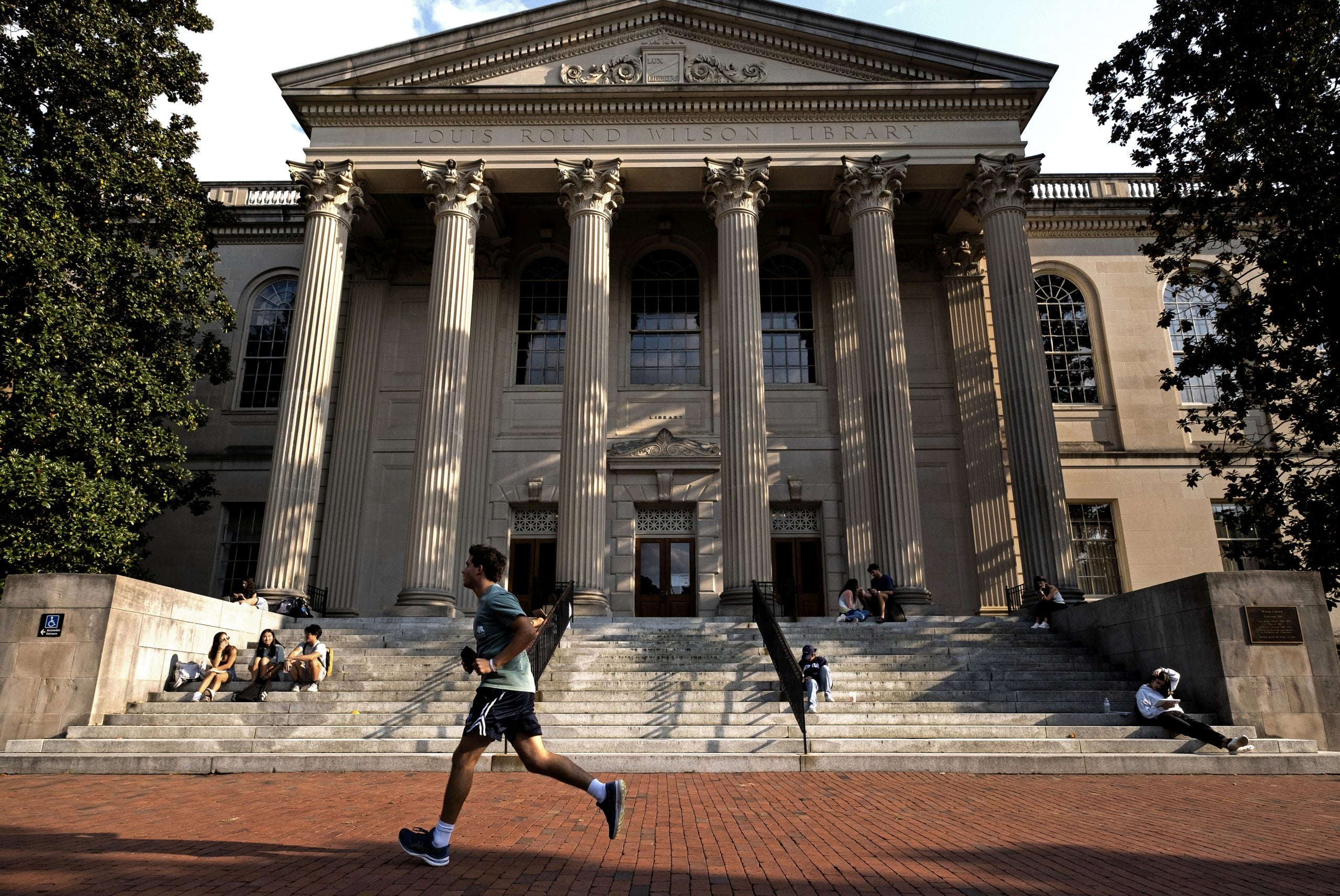 Several UNC-Chapel Hill students sitting on the steps of Wilson Library and a student jogging by in the foreground.