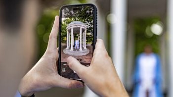 Close-up image of hands holding an iPhone to take a photo of a UNC-Chapel Hill graduate in Carolina Blue regalia posing in front of the Old Well.