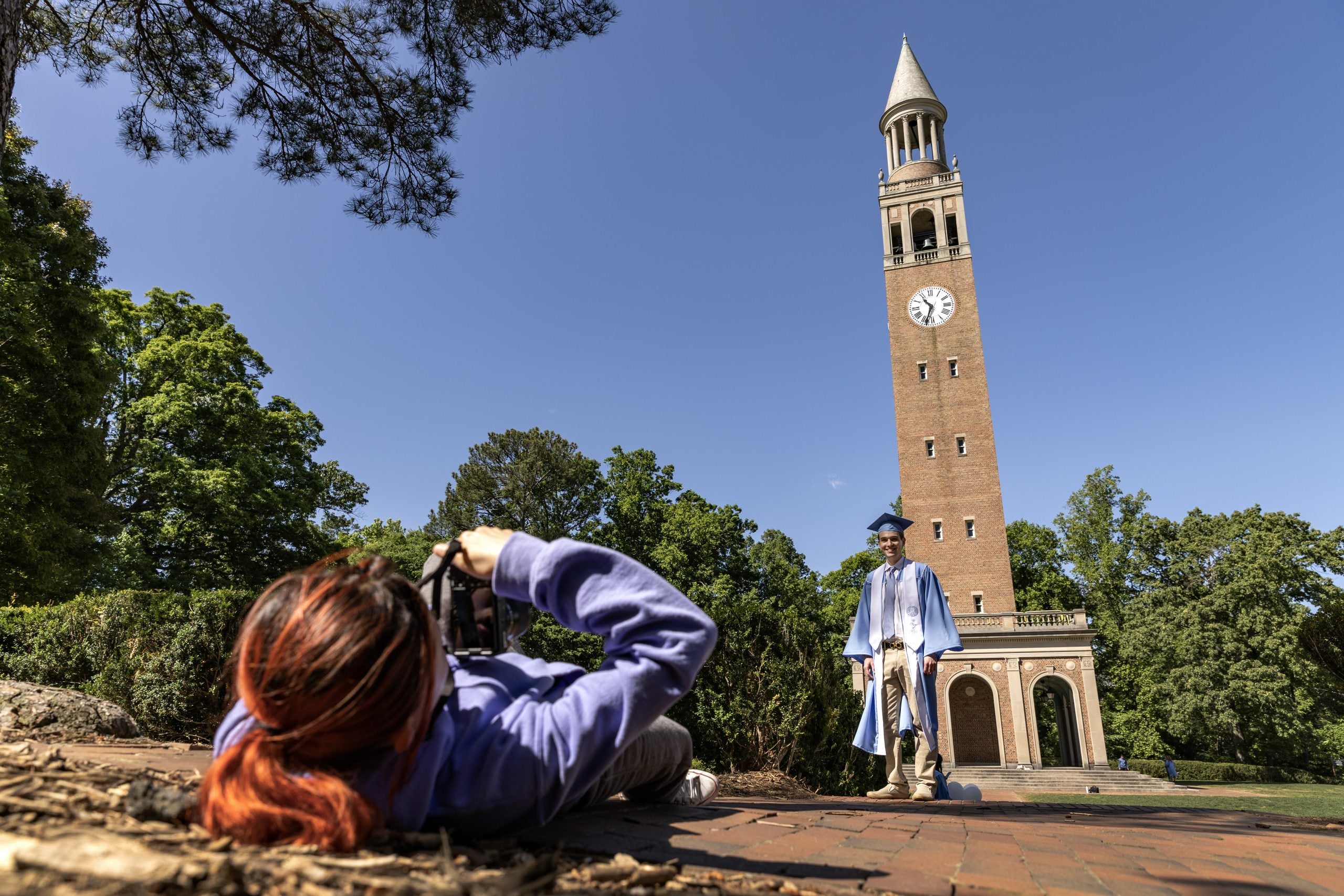 Person laying on the ground to make a photo of a student in Carolina Blue cap and gown with the Morehead-Patterson Bell Tower in the background.