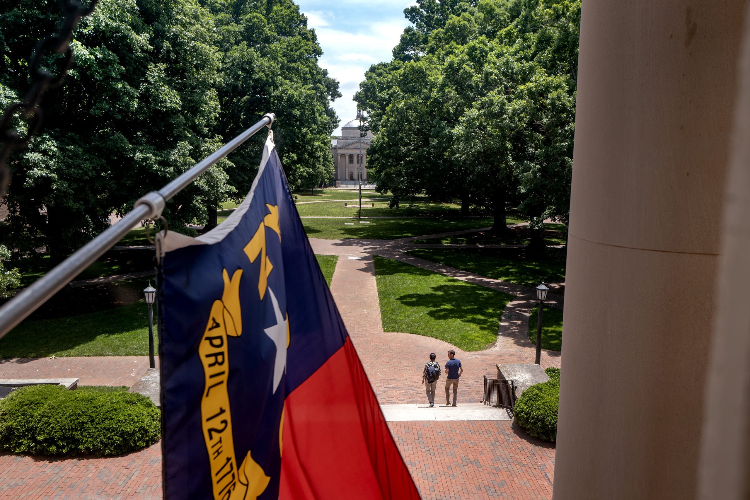State of North Carolina flag flying from South Building on the campus of UNC-Chapel Hill. Polk Place, with two people pictured, is seen in the background.