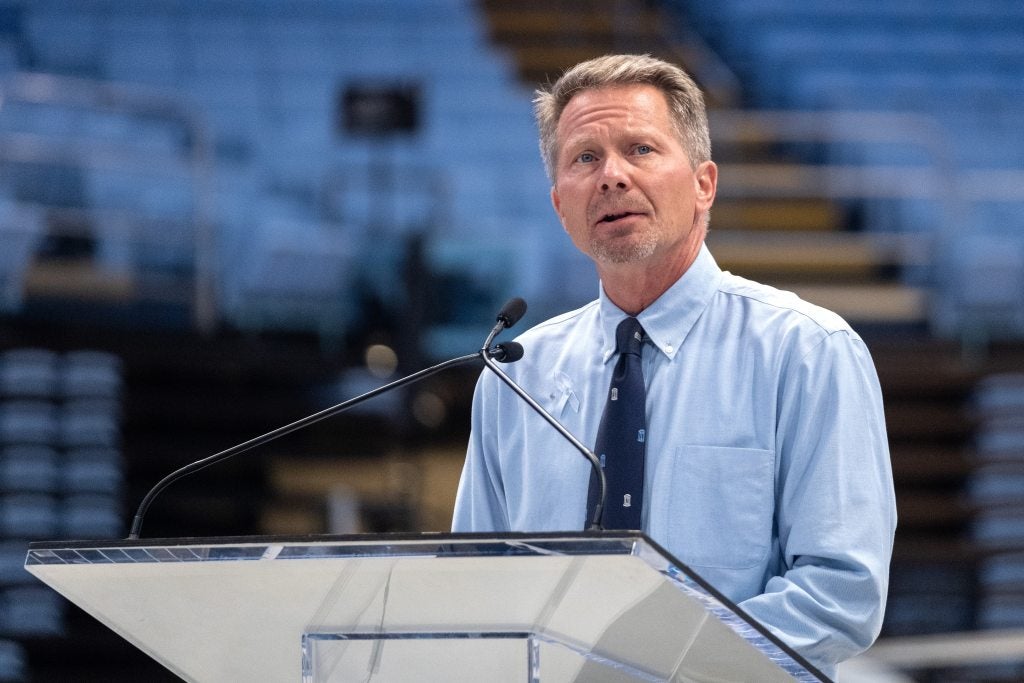 A man, UNC-Chapel Hill Chancellor Kevin M. Guskiewicz, speaking into a microphone at a podium in an arena while addressing a crowd at a vigil.