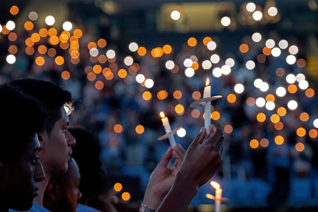 A large crowd of students, faculty, staff and community members standing and holding candles at a vigil in an arena.