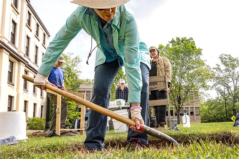 A woman with a shovel digging into grass in front of New East on the campus of UNC-Chapel Hill. Others look on in the background.