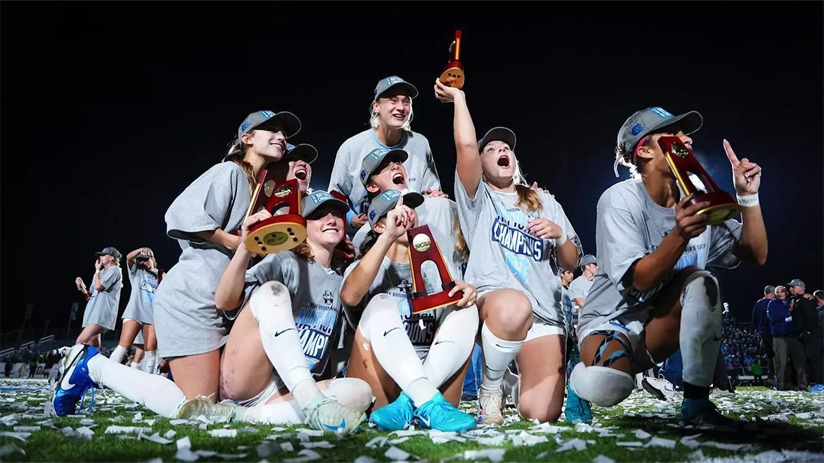 UNC women's soccer players celebrating as a group after national title on field. Players are wearing national championship T-shirts over their jerseys and hats, and one player is holding the trophy.