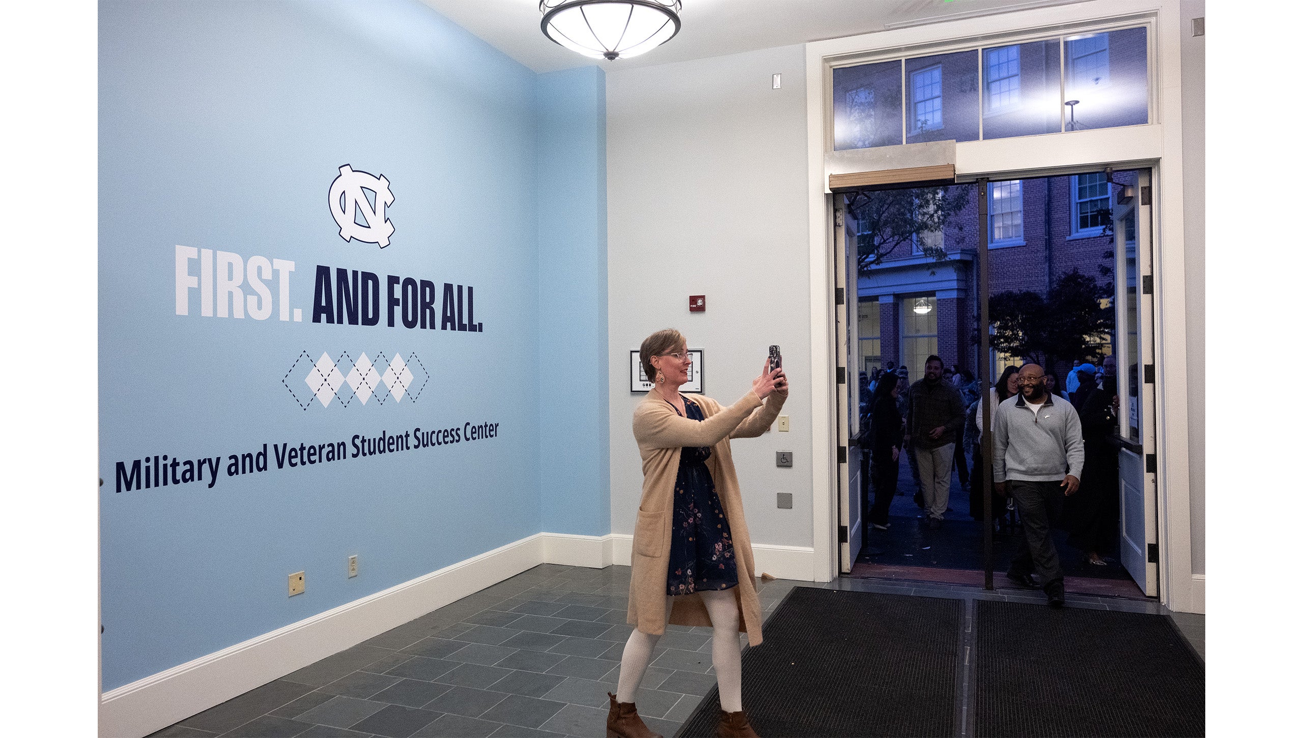 A woman taking a selfie inside the main entrance way o the Military and Veteran Student Success Center on the campus of UNC-Chapel Hill. Behind her is a Carolina Blue painted wall with the interlocking NC logo, an argyle pattern, text reading 