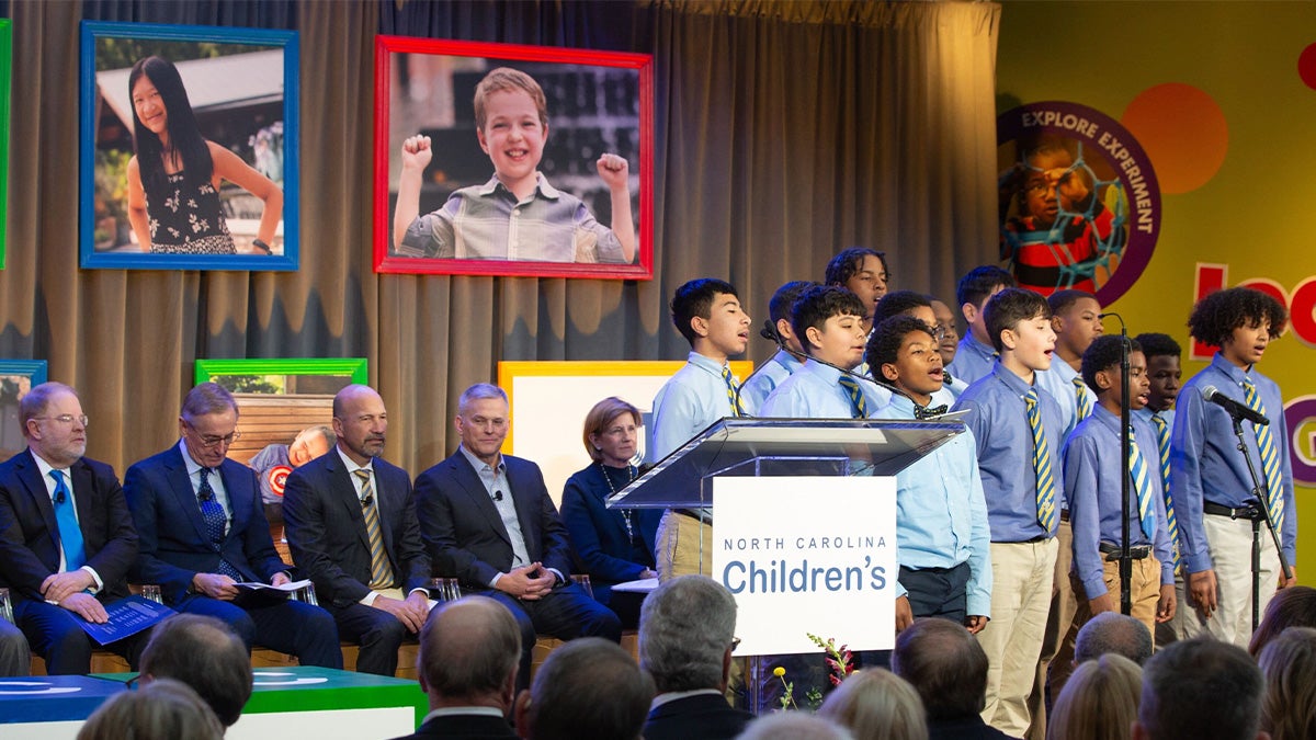A group of of children singing on stage with a group of administrators seated behind them at an event announcing a new children's hospital in North Carolina.
