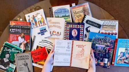 Hands holding open a vintage cookbook with a number of other cookbooks in the background.