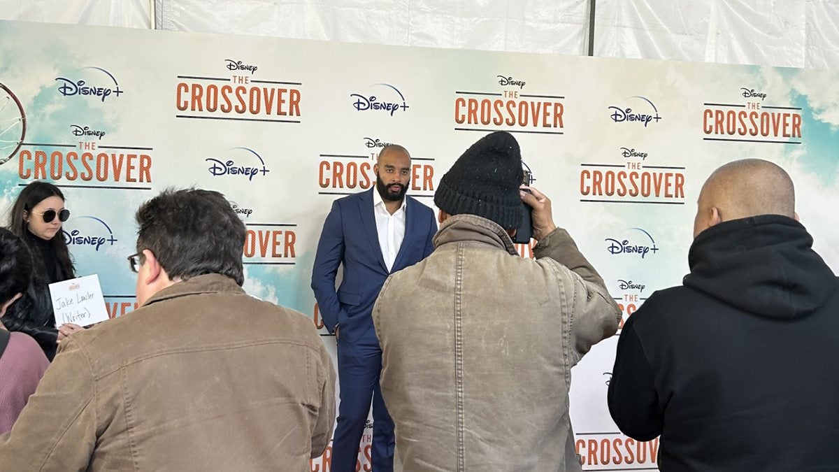 A man, Jake Lawler, wearing a suit and posing for a photo in front of a white backdrop with the Disney logo and the logo for a show called