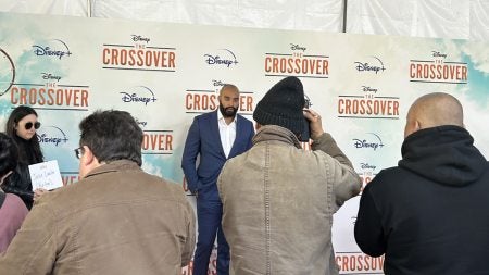 A man, Jake Lawler, wearing a suit and posing for a photo in front of a white backdrop with the Disney logo and the logo for a show called 