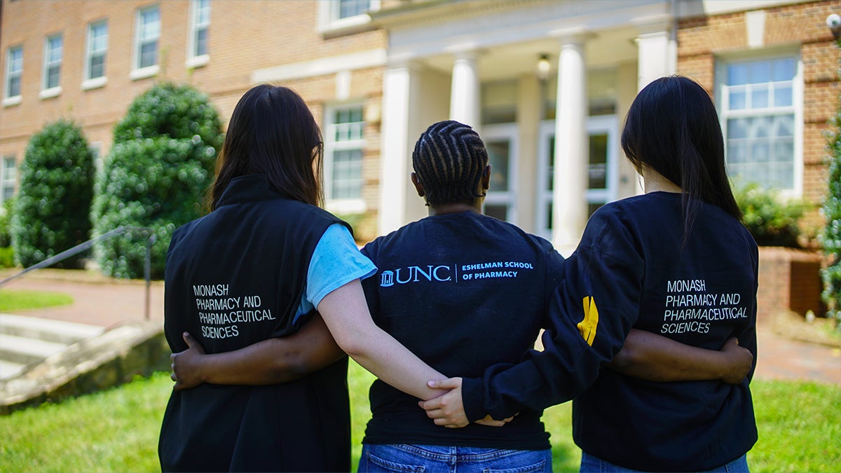 Three students stand together facing the Eshelman School of Pharmacy building. They are wearing T-shirts that say Eshelman or Monash University.