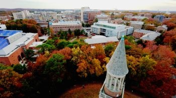 An aerial view of the Bell Tower and U.N.C. Chapel Hill's campus in the fall. The trees have orange and yellow leaves.