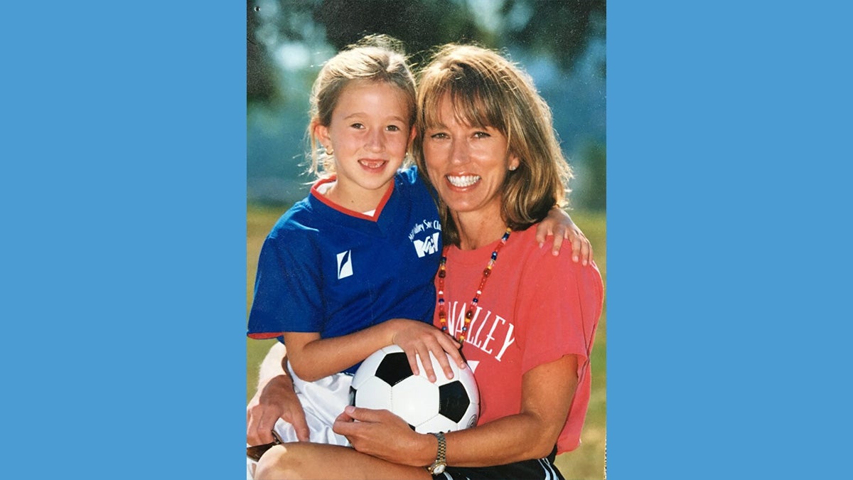 Old soccer photo of a young Darcy McFarlane holding soccer ball and posing with her mother.