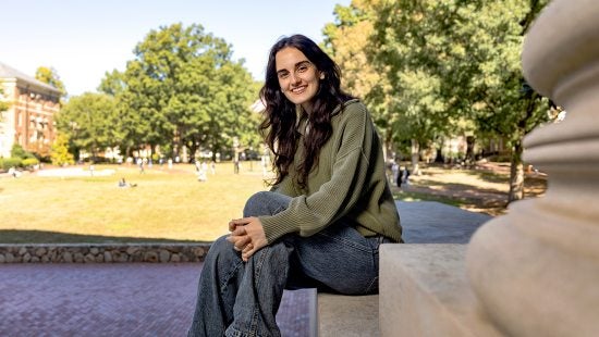 Gunes Tugcu sitting on a stone ledge outdoors on a sunny day, smiling at the camera. They are wearing a green sweater and jeans, with a grassy campus quad and trees in the background.