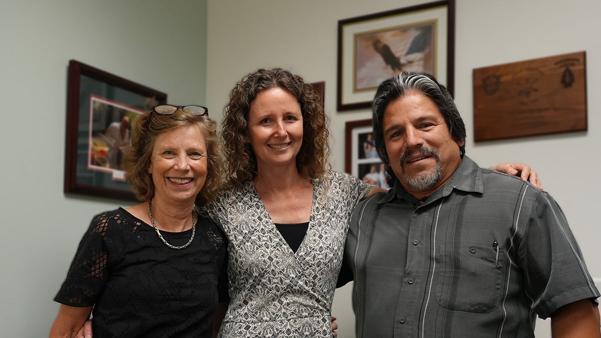 Three people stand arm in arm posing. One woman has blonde hair and a black shirt, next to another woman with brown curly hair and a white top. The third individual is a man wearing a gray shirt and sporting a beard.