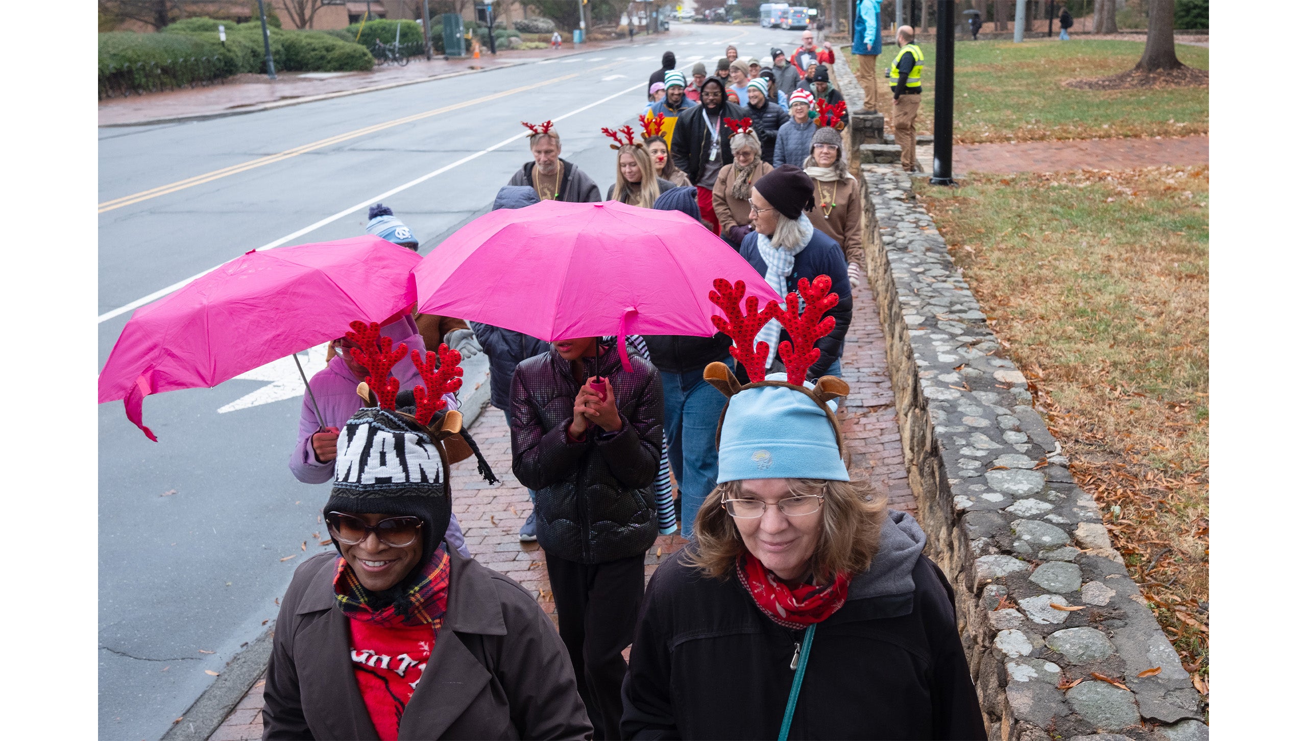 Long line of UNC-Chapel Hill employees in festive holiday costumes, and some with umbrellas, walking down a brick sidewalk aside South Road during the Jingle Bell Jog.