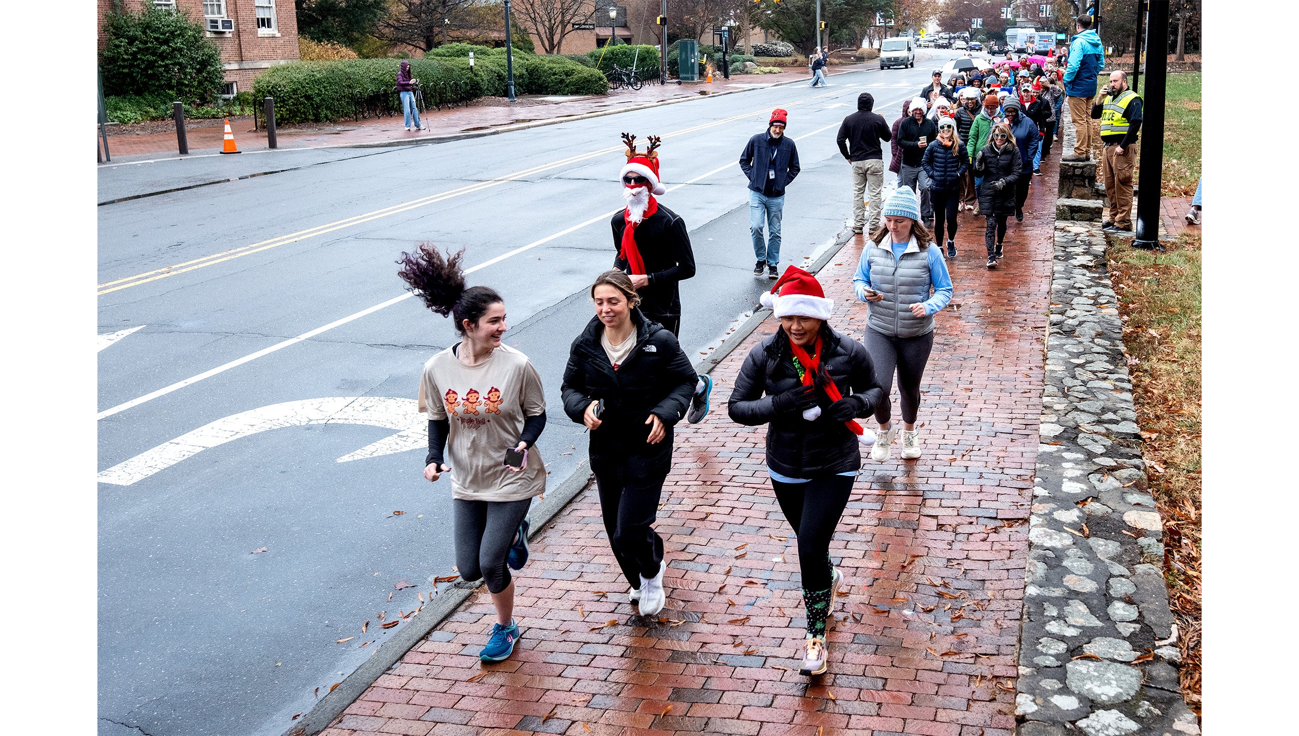 Five UNC-Chapel Hill employees jogging during the Jingle Bell Jog down a brick sidewalk aside South Road ahead of a crowd of walkers.