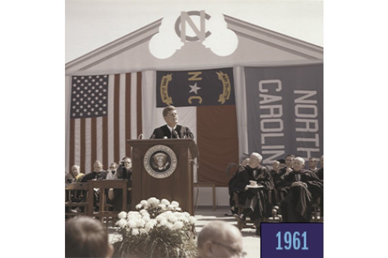 A man, former president John F. Kennedy, speaking at a podium on an outdoor stage on the campus of UNC-Chapel Hill. A few rows of people are sitting behind him, and there is a backdrop featuring the American flag, the North Carolina state flag and a Carolina Blue flag reading 