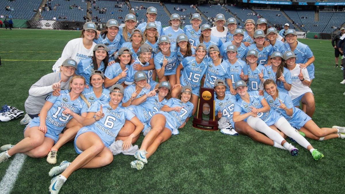 UNC women's lacrosse team celebrating and taking team photo on field after game with national title trophy.