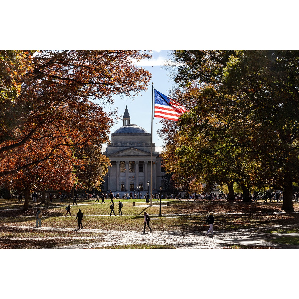 Polk Place on the campus of U.N.C. Chapel Hill on a sunny fall day. People are seen walking on brick pathways and sitting on the lawn, and a large American flag is flying. Wilson Library is seen in the background.