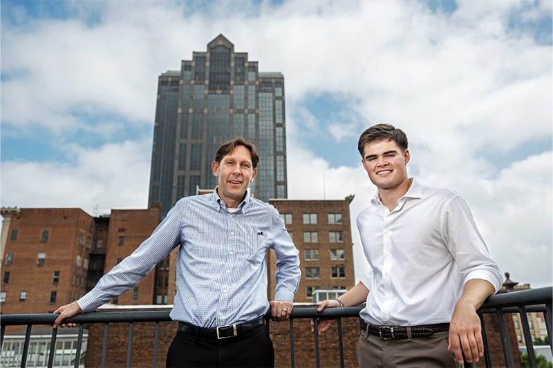 Andrew Stewart and Reece Hawk posing for a portrait while leaning back against a railing on a rooftop balcony in downtown Raleigh. Other tall downtown buildings are seen in the background.