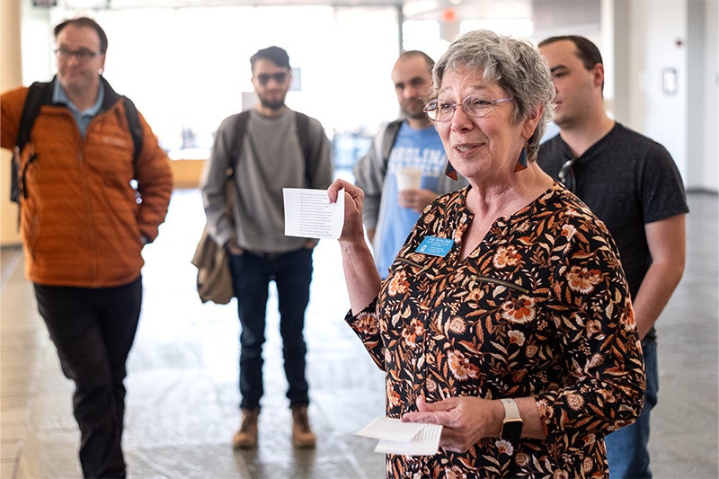 A woman, Gigi Taylor, holding up a slip of paper with various conversation-starter questions on it at the Speaking Group's weekly meetup. Four participants are seen in the background behind her.