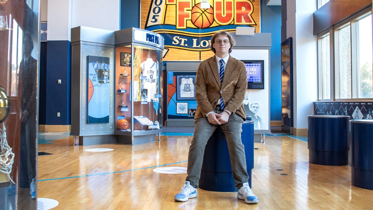 Connor Kerr posing in front of sports memorabilia wearing a dark tan suit.