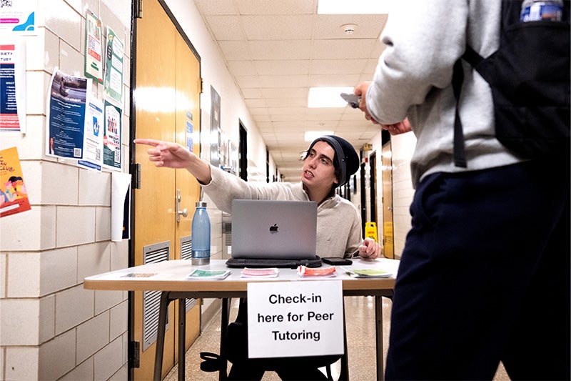 A student sitting at a check-in desk in a hallway directs a fellow student toward a room by pointing to his right.