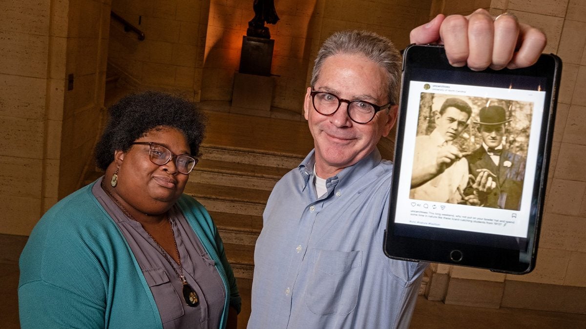 Lolita Rowe and Nick Graham, UNC-Chapel Hill archivists, posing for a photo in the lobby of Wilson Library. Graham is holding up an iPad displaying an Instagram post from the uncarchives account, which he and Rowe run.