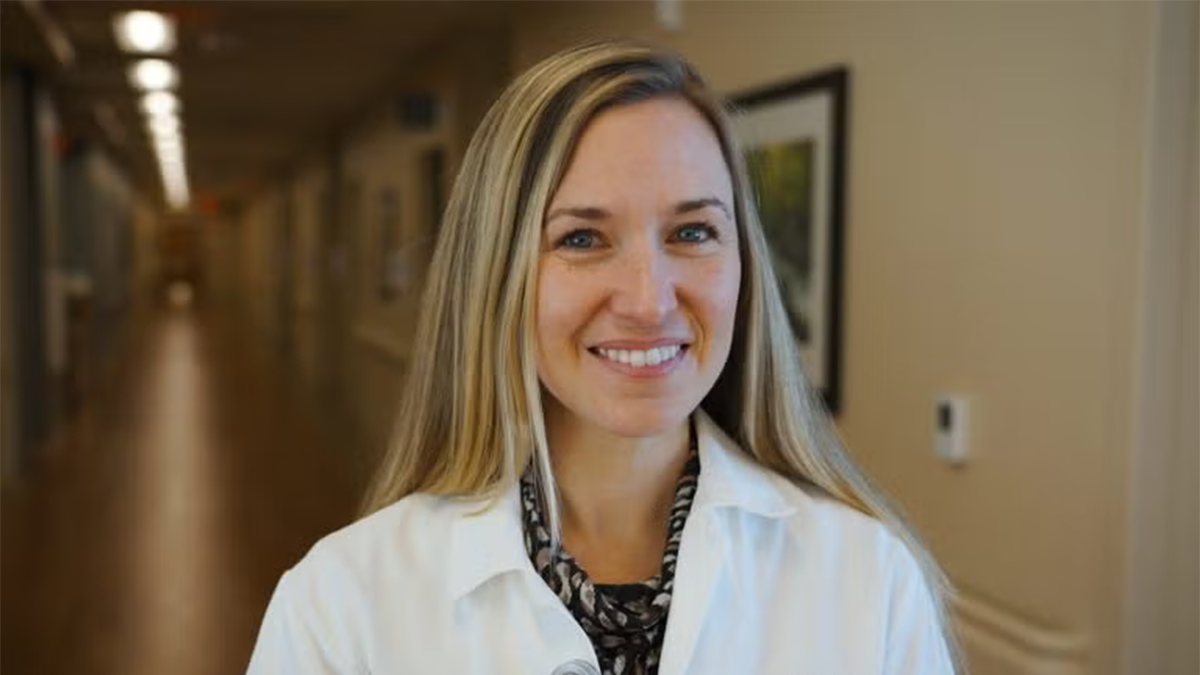 A smiling woman wearing a white medical coat stands in a softly lit hallway. She has long, straight blonde hair and is looking directly at the camera. The background shows a corridor with framed artwork on the walls.