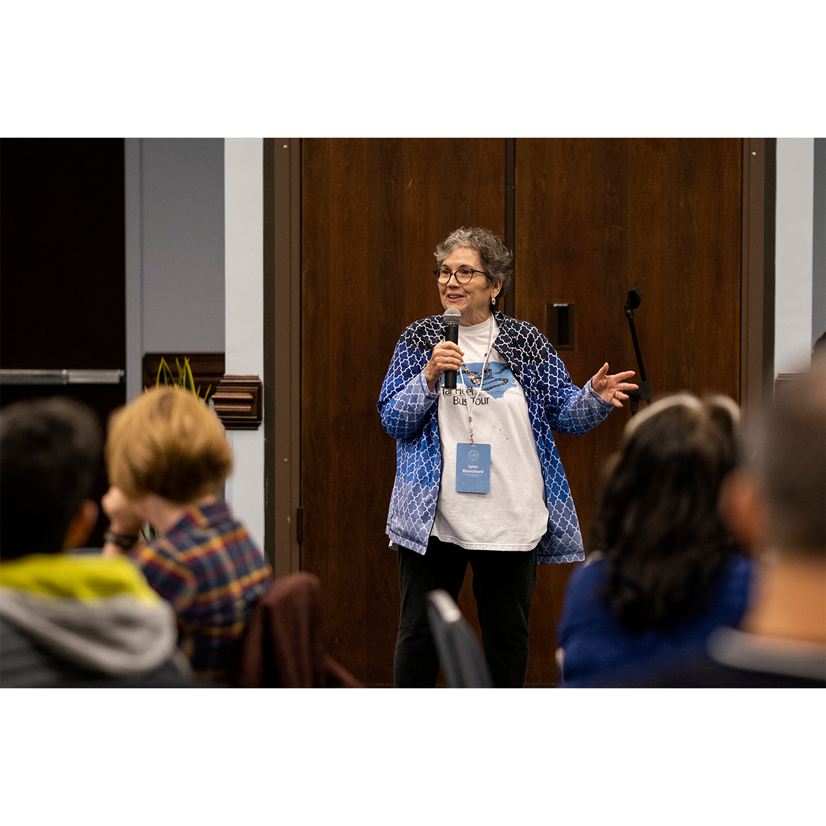 A woman, named Lynn Blanchard, holding a microphone and speaking to a group of bus tour attendees in an indoor meeting room.