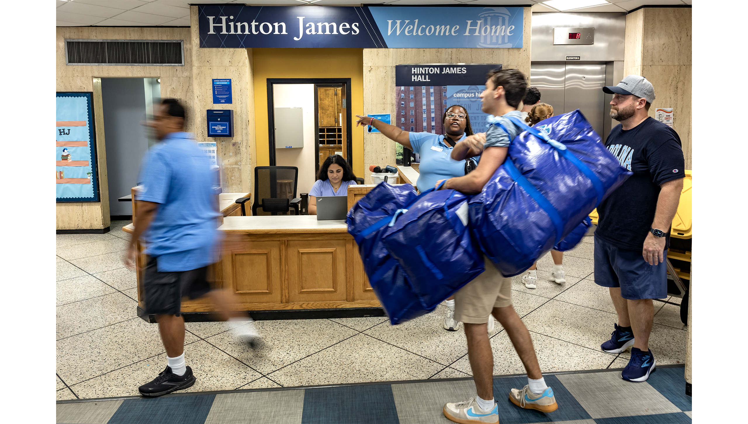 Student carrying large blue bags with family members walking around as a staff member points them in the direction they need to go.
