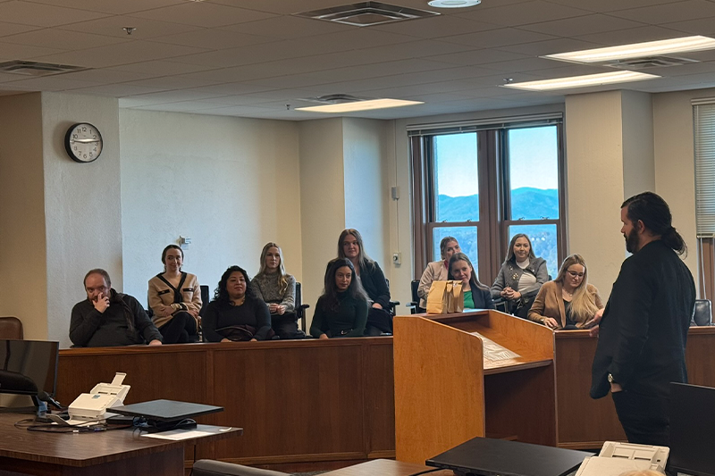 A man speaks to a group of people seated in a wooden room with large windows overlooking a mountain view.